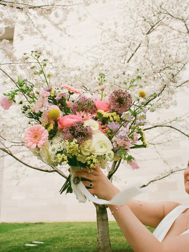 Bride holds spring pastel bouquet up to cherry blossom tree.