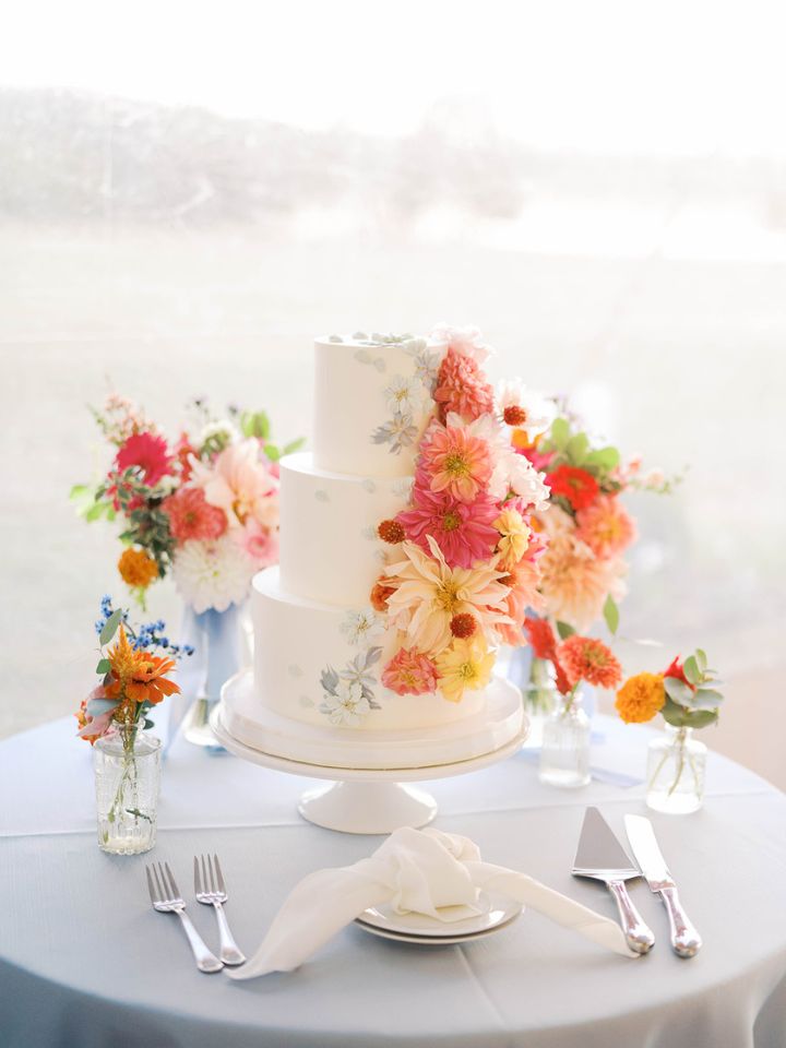 A classic white three tier wedding cake on a pedestal stand sits on a table with light blue cloth, floral bud vases, and is decorated in colorful dahlia flowers.