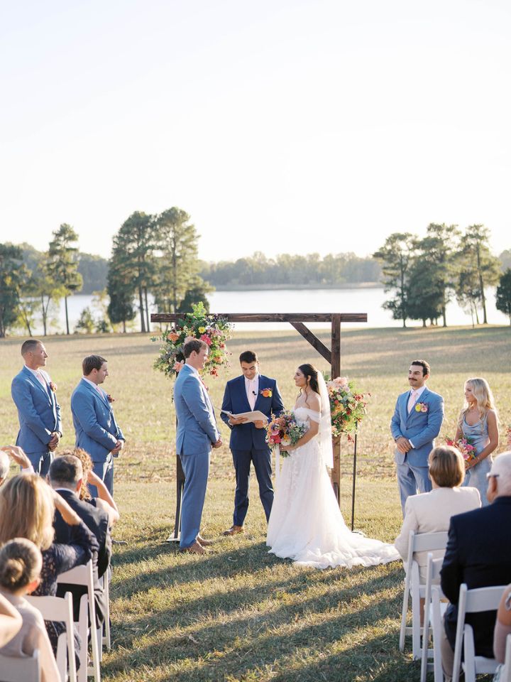 Bride and groom stand at wooden arch in field for outdoor wedding ceremony.