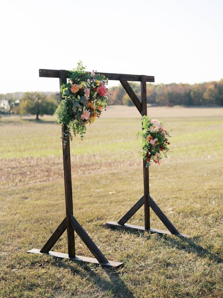 Wooden arch for wedding ceremony stands outdoors, with two flower arrangements on it.