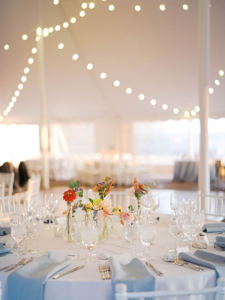View inside a white reception tent with bistro lighting, white tablecloths with blue napkins, and colorful fall flowers in bud vases.