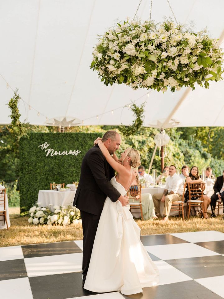 Couple dances under large green and white flower chandelier in wedding reception tent.