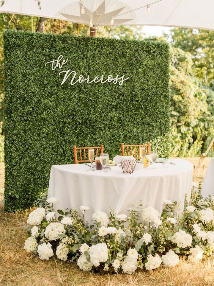 Large green boxwood wall with last name, behind sweetheart table with white flowers surrounding the base.