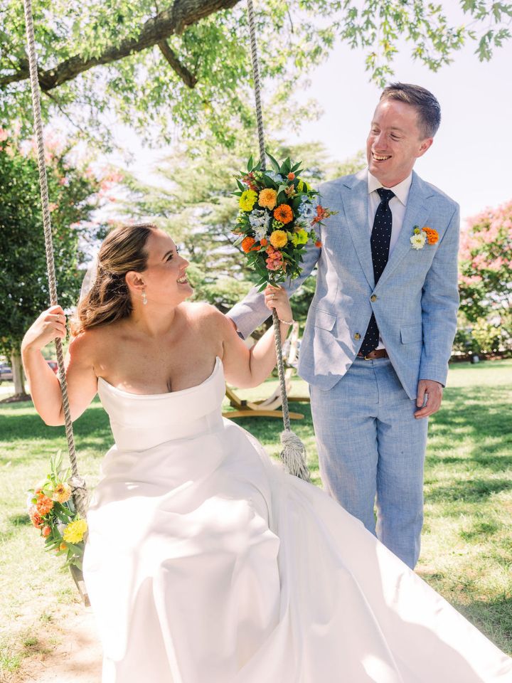 swing with flowers Bride sits on swing that is decorated in flowers, smiling at groom standing beside her.