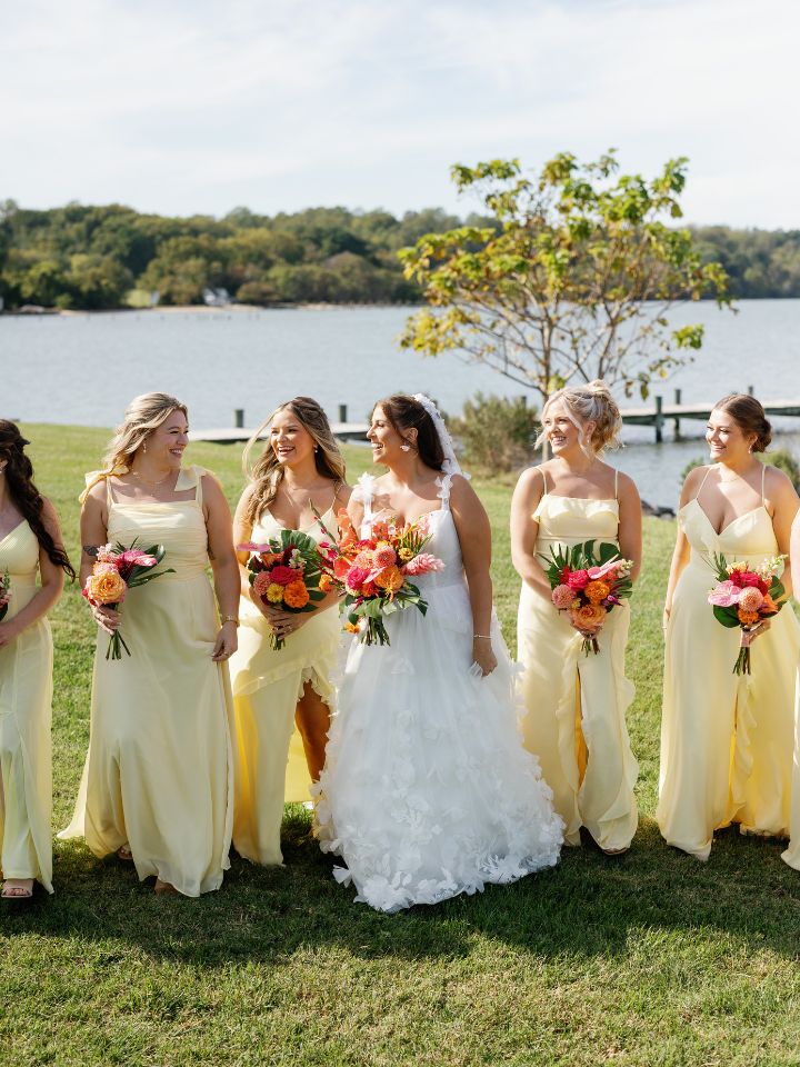 Bride walks with bridesmaids in butter yellow dresses, holding flowers.