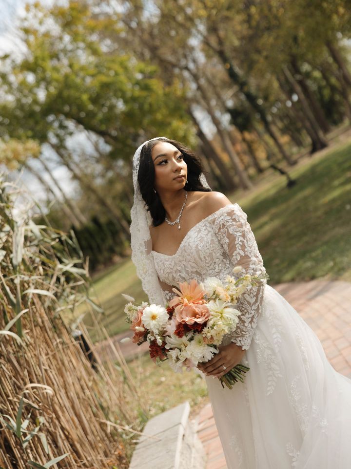 Bride poses with lace sleeves and veil in front of grasses, holding her fall bouquet.