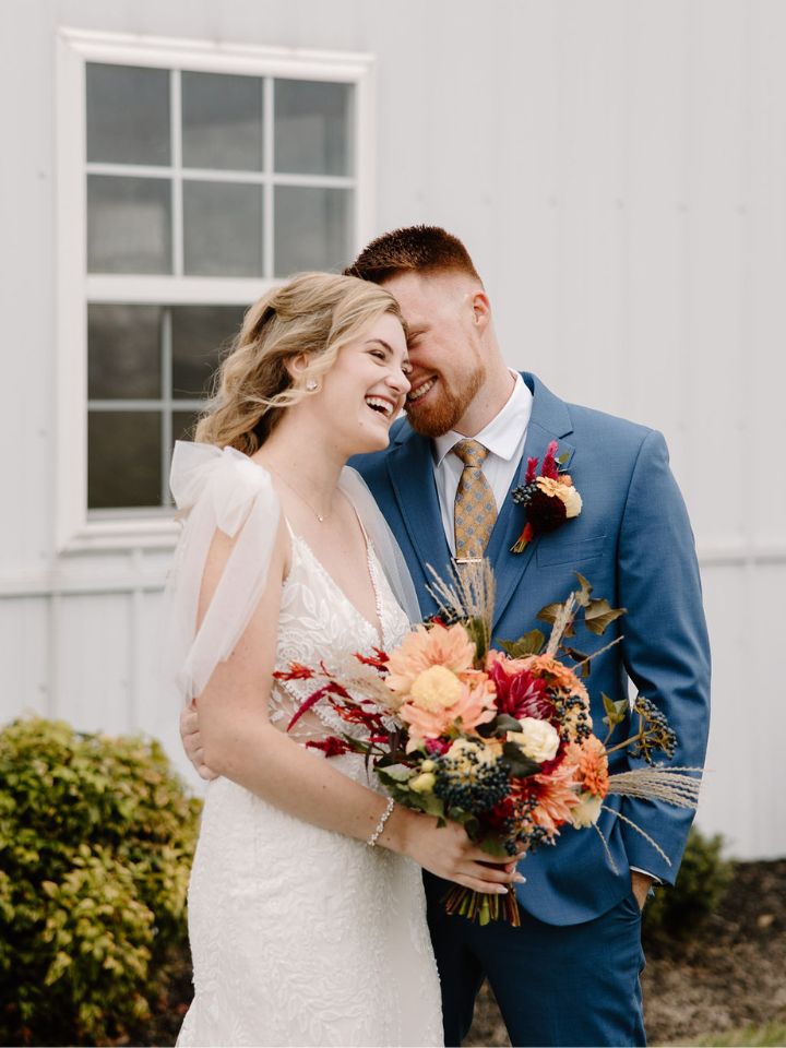 Bride holds her fall wedding bouquet and smiles with groom in blue suit.