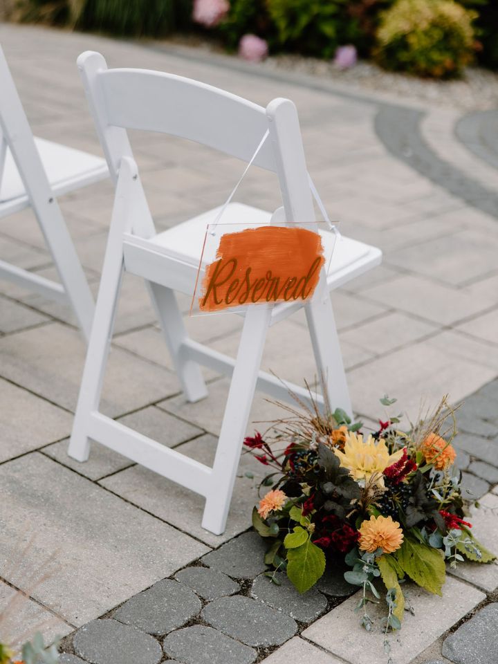 White chair at wedding ceremony aisle with orange reserved sign and flower arrangement at base.