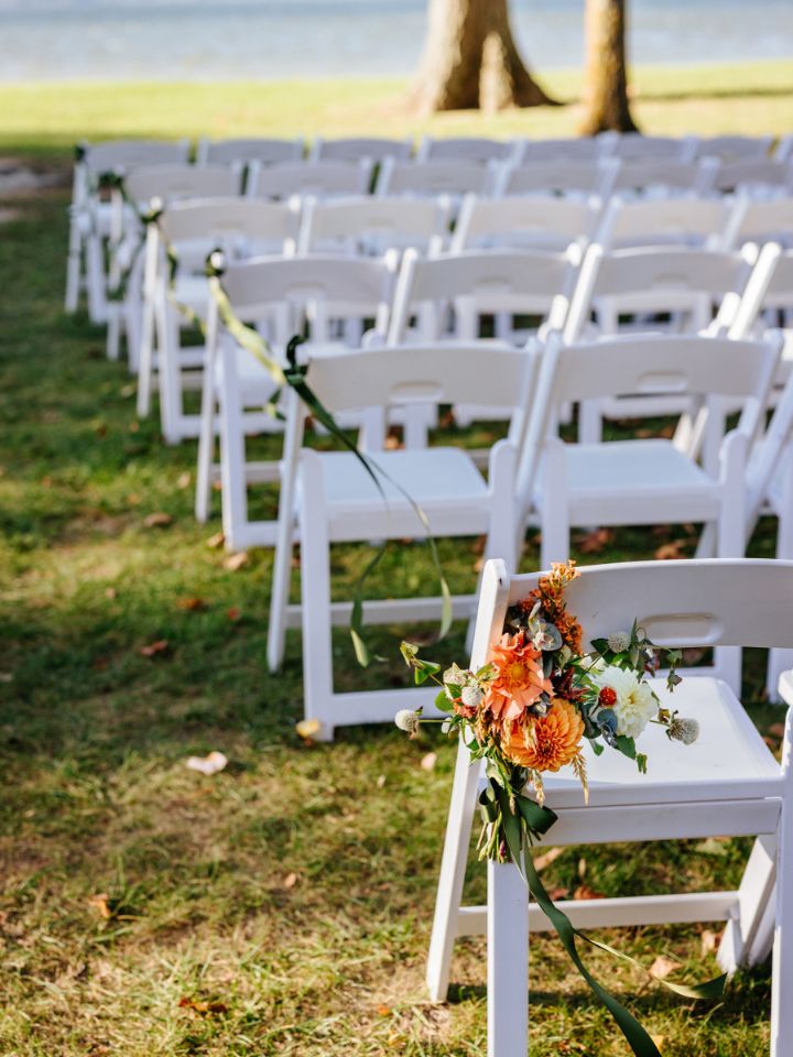 White folding chairs at wedding ceremony with flowers and olive green ribbons tied to sides.