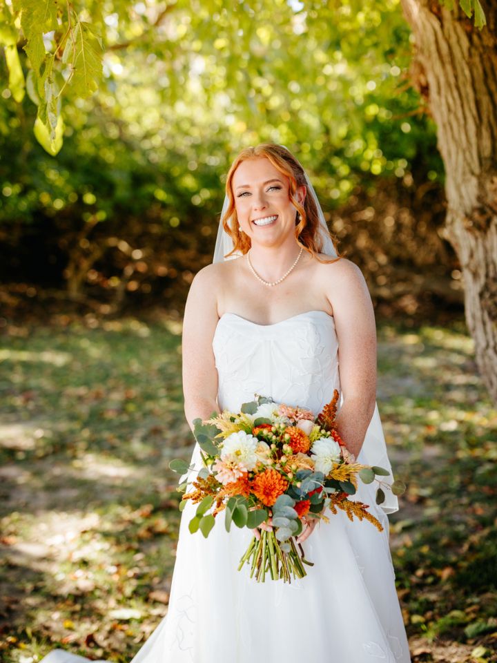 Bride poses under tree holding her fall flower bouquet.