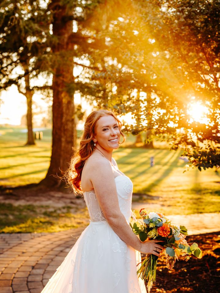 Bride poses under trees at golden hour, holding her fall flower bouquet.