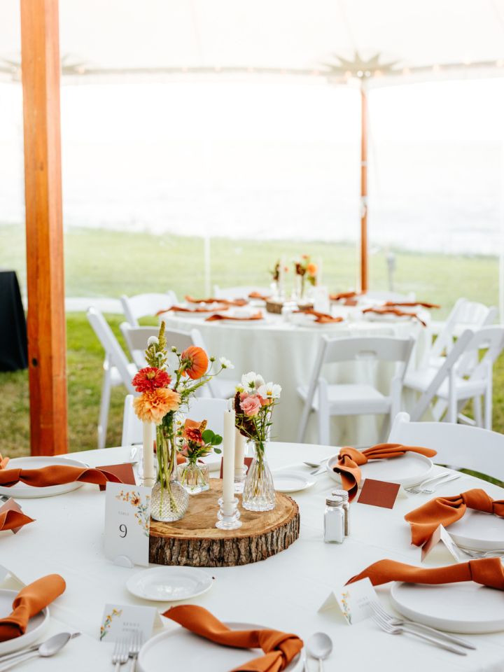 Inside reception tent at wedding with white linens, rust red napkins, and centerpiece with wood slab, candles, and bud vases.