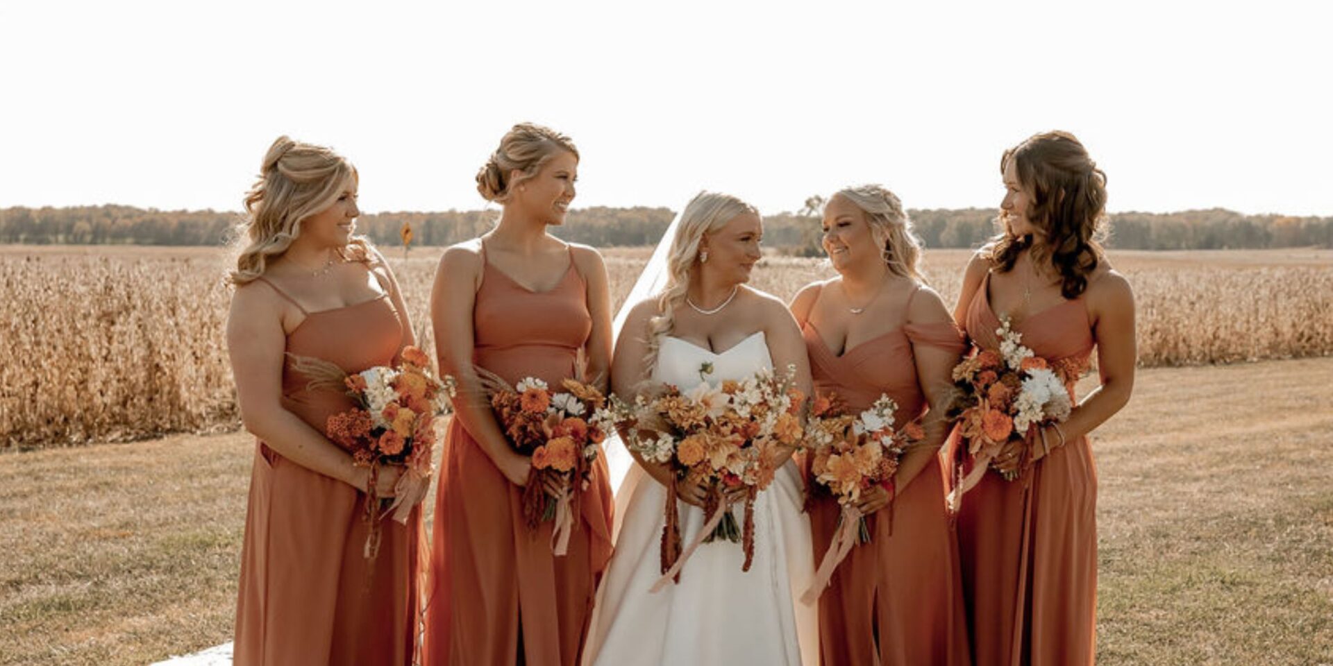 Bride and bridesmaids in rust orange stand next to wheat field with flower bouquets.