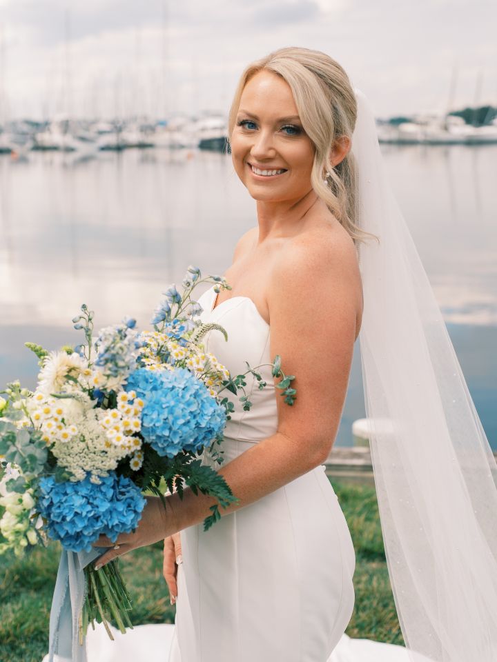 Bride smiles at camera, holding her summer blue and white bouquet.