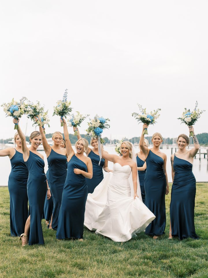 Bridesmaids in navy blue dresses hold up bouquets in air.