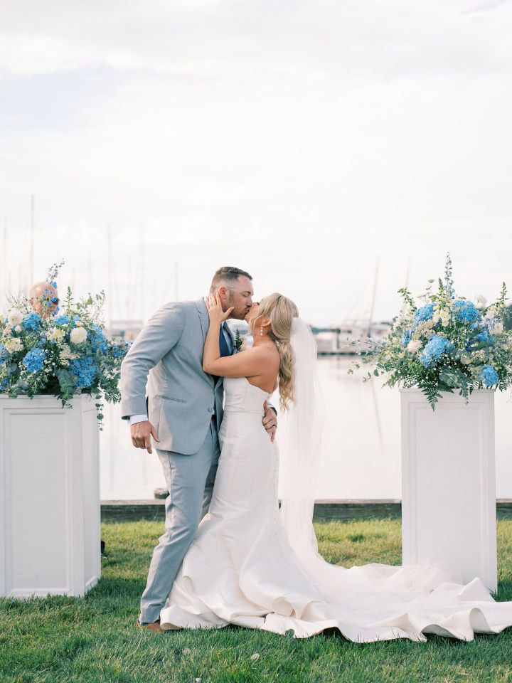 Bride and groom kiss between white pedestals with flower arrangements on top.