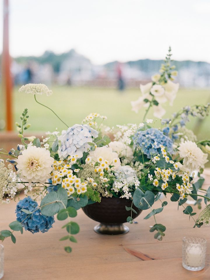 Hammered copper compote dish with blue and white summer wedding flower arrangement sits on a wooden harvest style table.