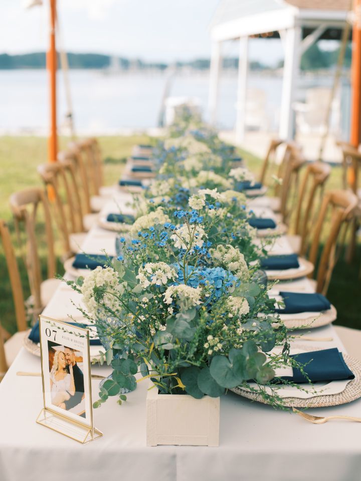 White planter style box with blue and white flowers runs end-to-end on a long wedding reception table.