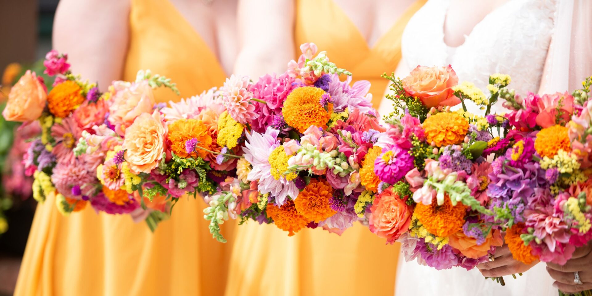 Closeup of bridesmaid in gold and bride in a line, holding colorful summer bouquets.
