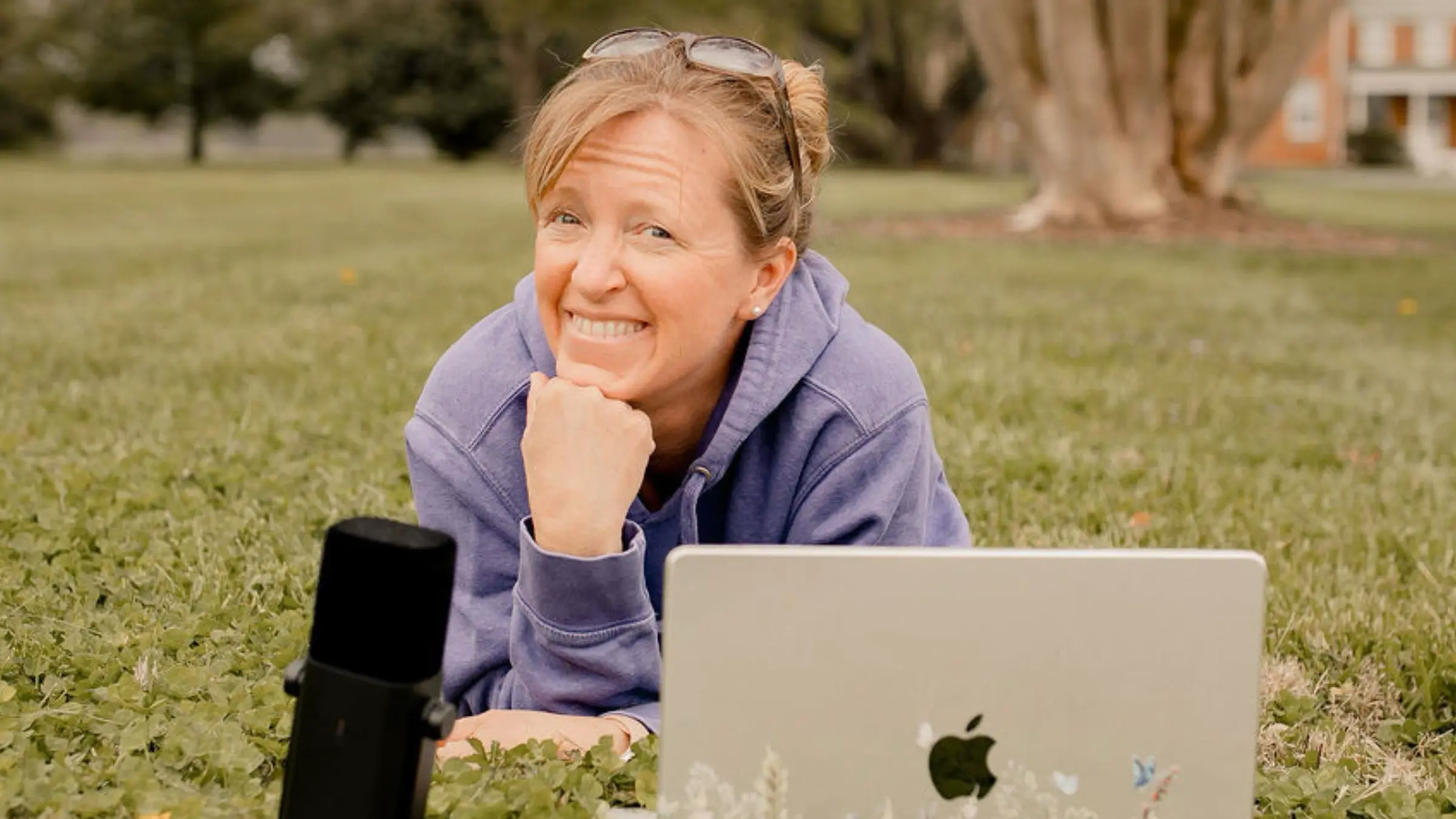 Liza smiling with her laptop and her podcast microphone in the grass