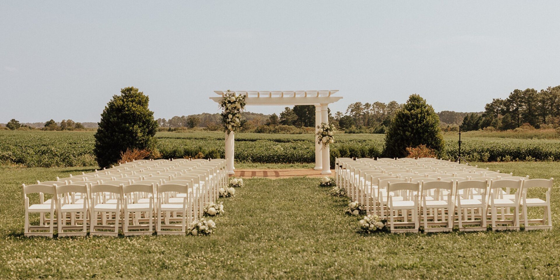 View of a wedding ceremony at a white pergola with rows of white padded folding chairs.