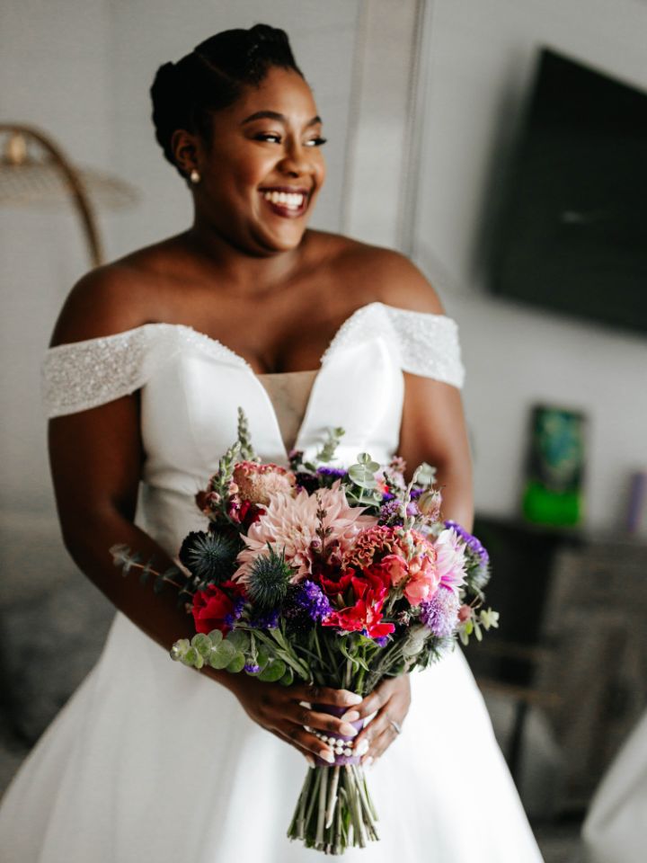 Bride smiles while holding her purple, magenta, and blue bouquet.