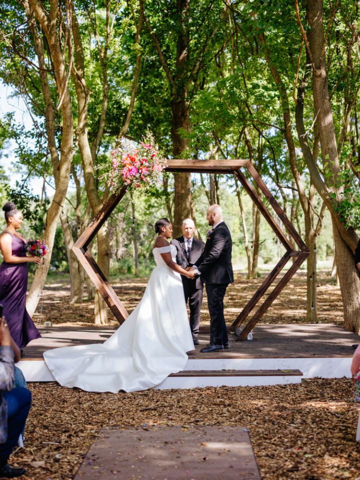 Bride and groom stand under hexagon arch for wedding ceremony in the woods.