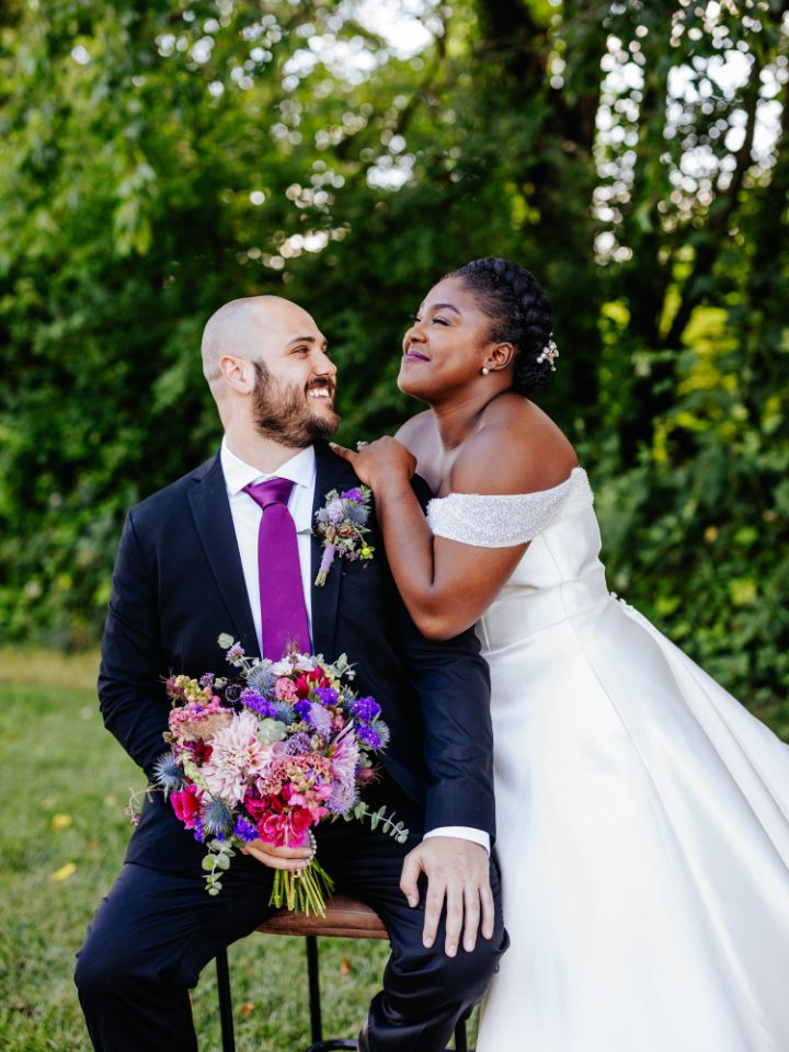 Groom sits on chair, bride stands at shoulder.