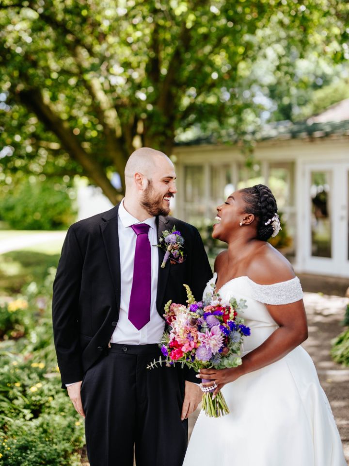 Bride and groom smile at each other, him in black suit and purple tie, her holding bridal bouquet and wearing white dress.