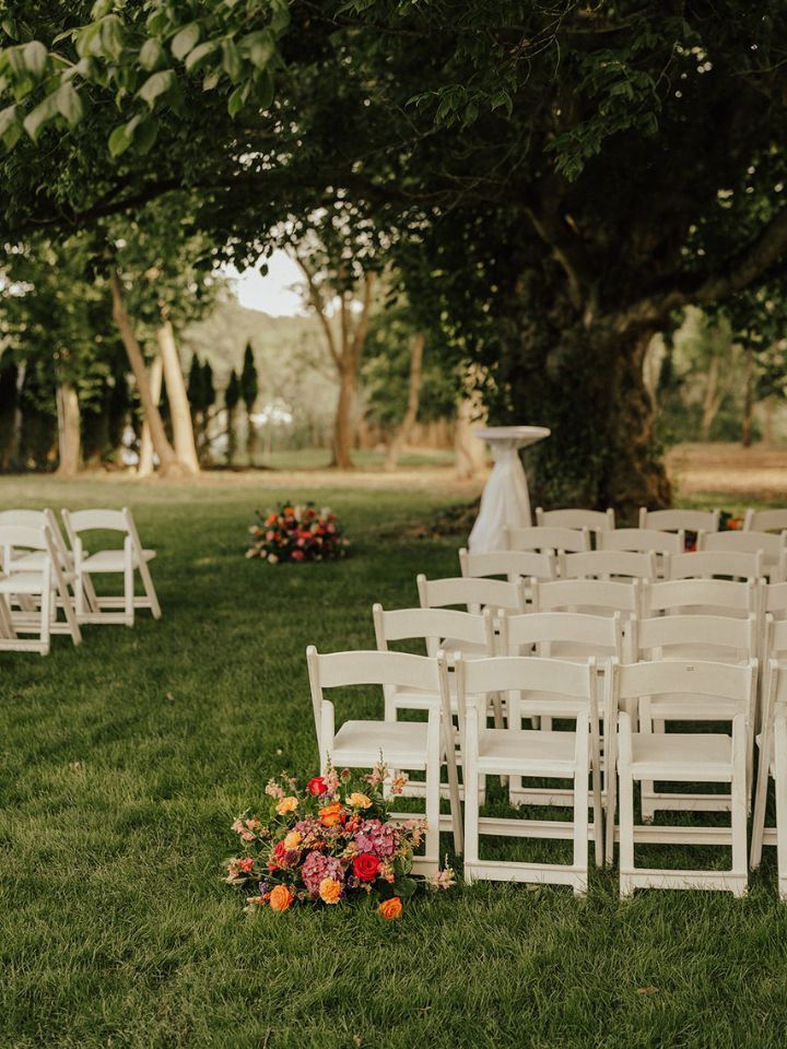 View of outdoor wedding ceremony under large tree in grass, with white folding chairs, and large flower arrangements.