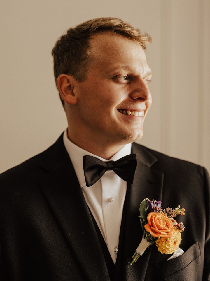Groom looks off camera, wearing black tuxedo and colorful boutonniere.