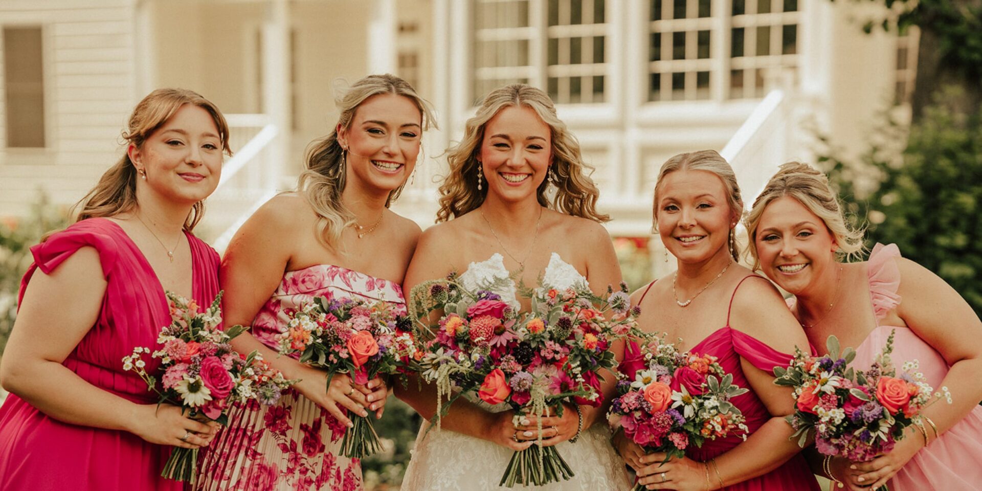 Bride and bridesmaids in a line, wearing pink dresses, holding flower bouquets.