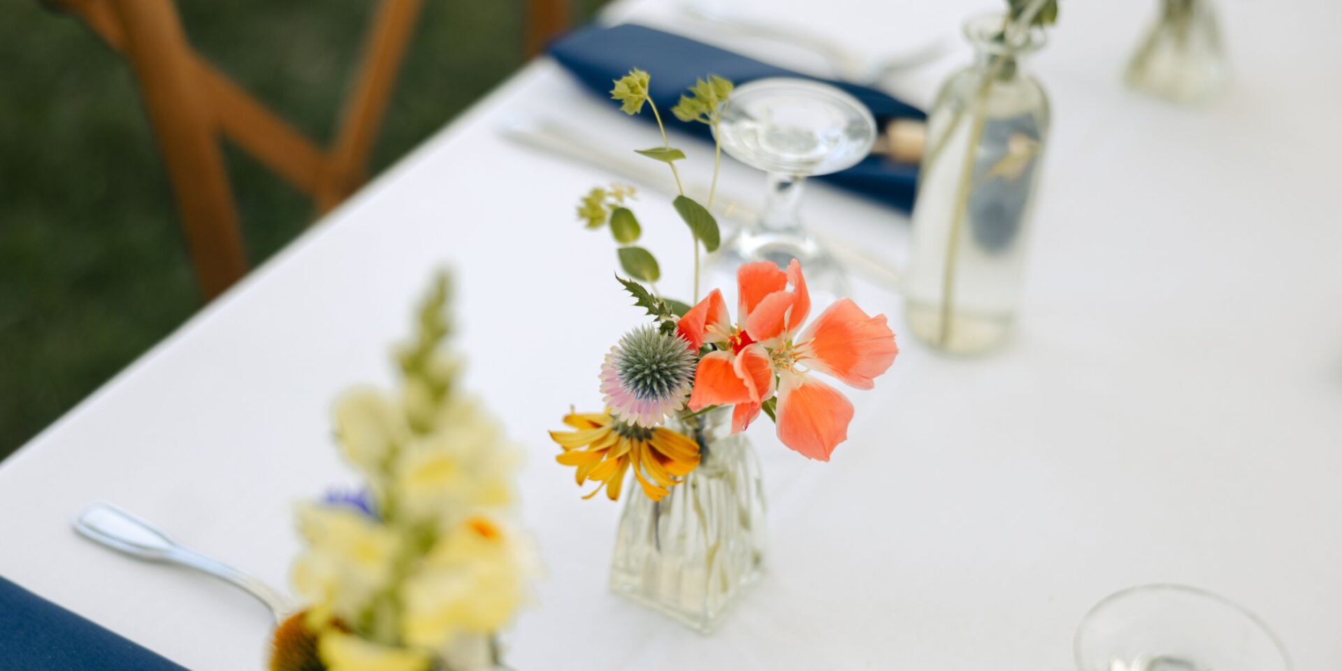 Simple glass bud vases on white table linen with colorful summer flowers.