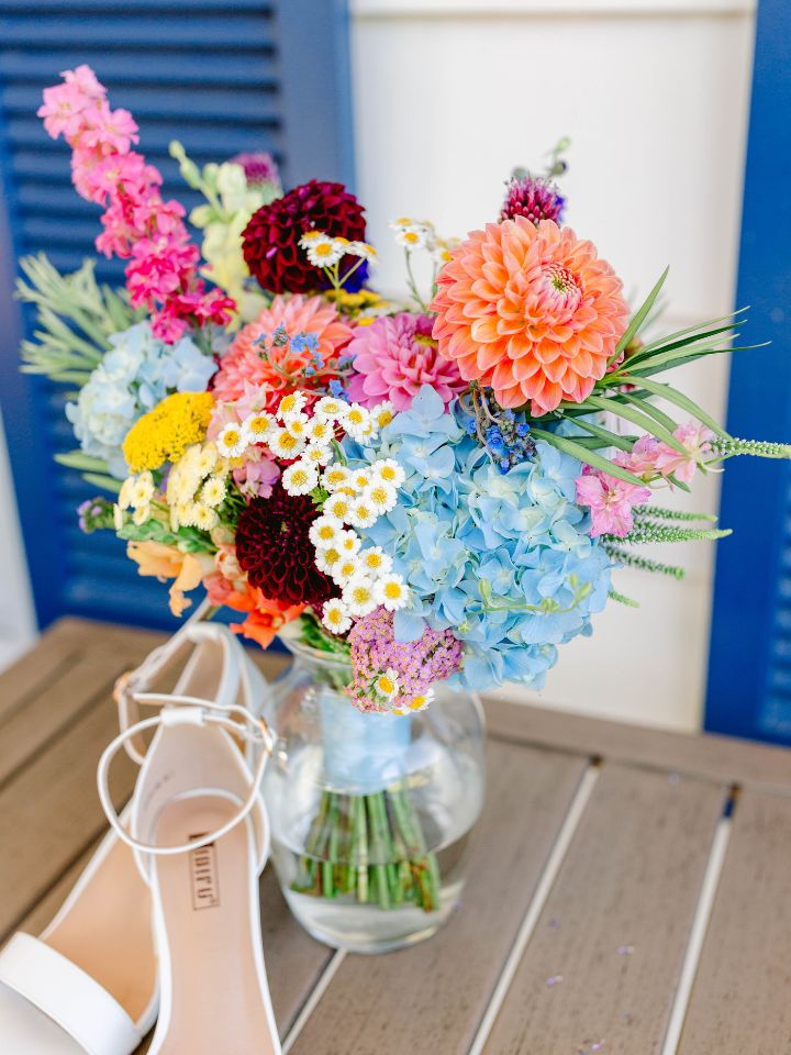Bridal bouquet rests in a vase, with white shoes, on brick step and blue shutters in background.