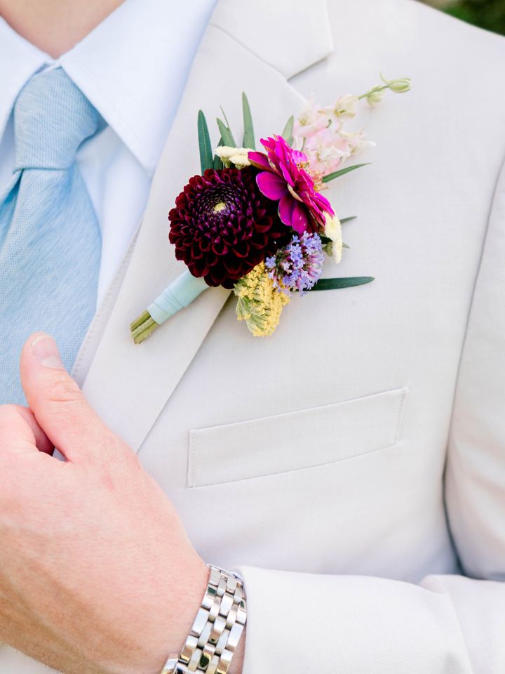 Closeup of groom's boutonniere with burgundy dahlia on a tan suit with blue tie.