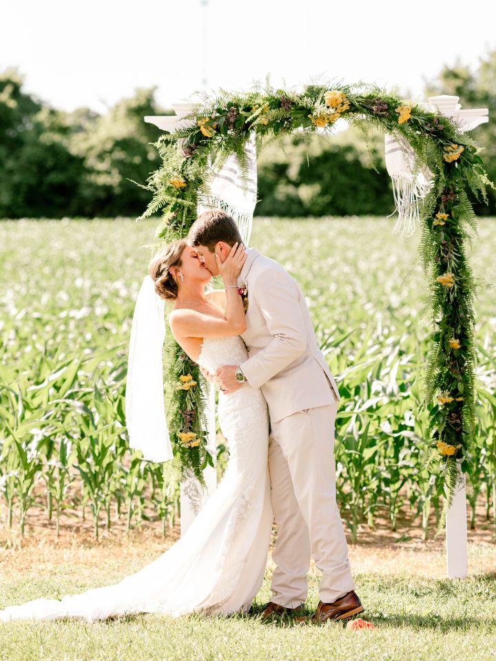 Bride and groom kiss at outdoor wedding ceremony with garland on small arbor.