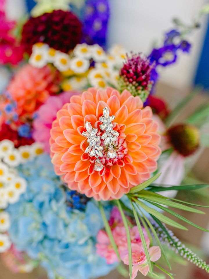 Earrings sit on top of bright orange dahlia flower, with blue hydrangea and other flowers in back.