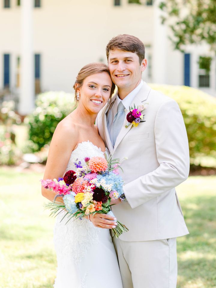 Bride and groom pose for photo with large manor style house in background.
