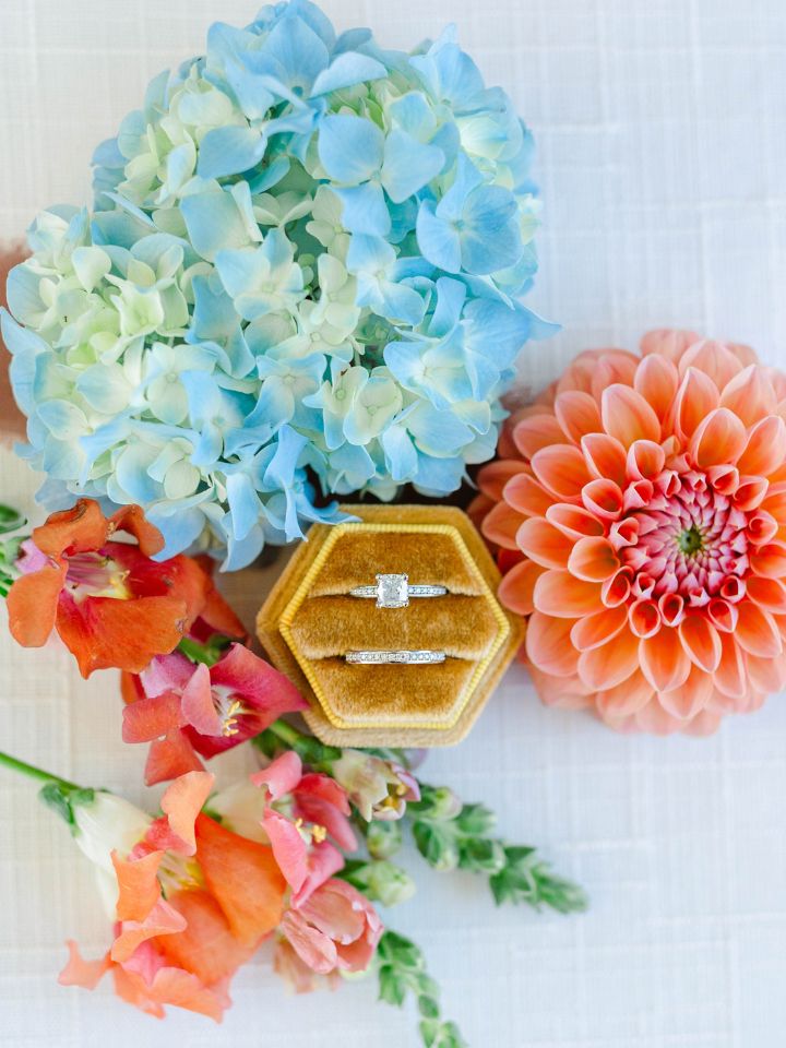 Mustard yellow velvet ring box with wedding rings sits with vibrant summer flowers.