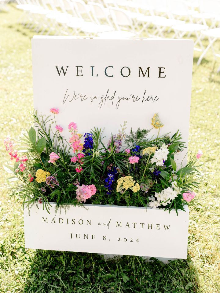 White standing welcome sign with window box style front and flower arrangement.