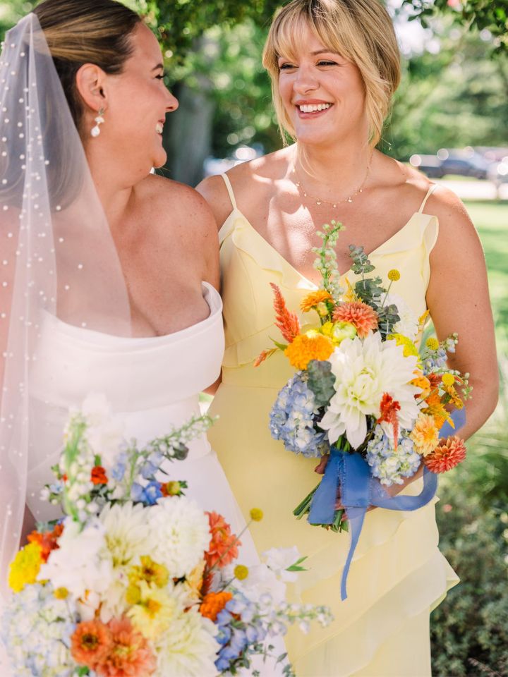 Bride and bridesmaid in butter yellow dress smile at each other, holding flower bouquets.
