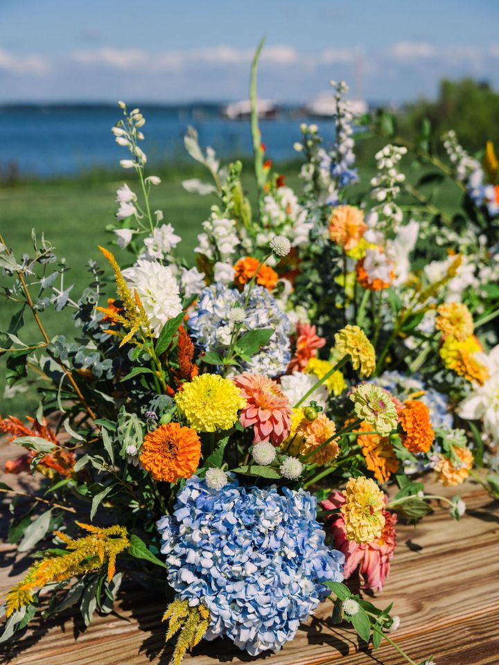 Large flower arrangement in blue, white, yellow, and orange at an outdoor waterfront wedding ceremony.