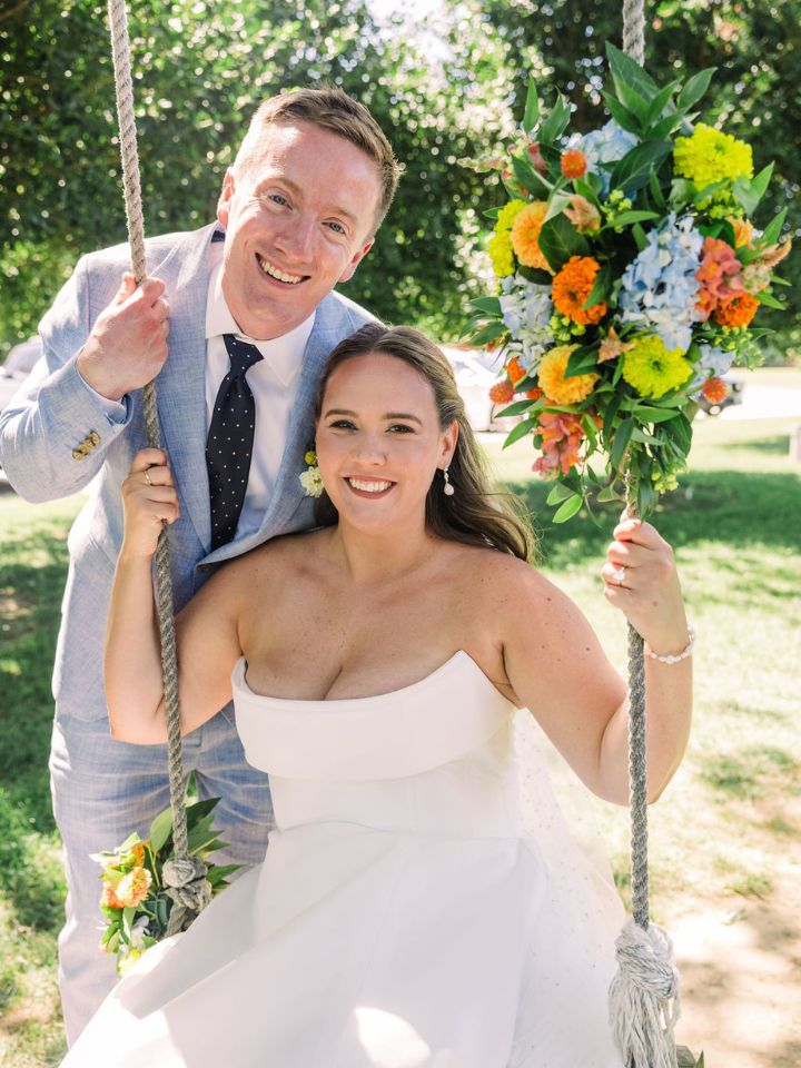 Bride sits on swing decorated with flowers, with groom standing behind her.