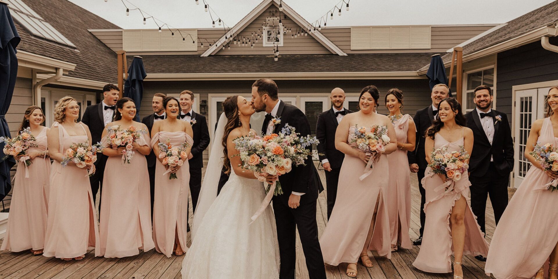 Bride and groom kiss in front of wedding party in black tuxes and soft pink bridesmaid dresses.