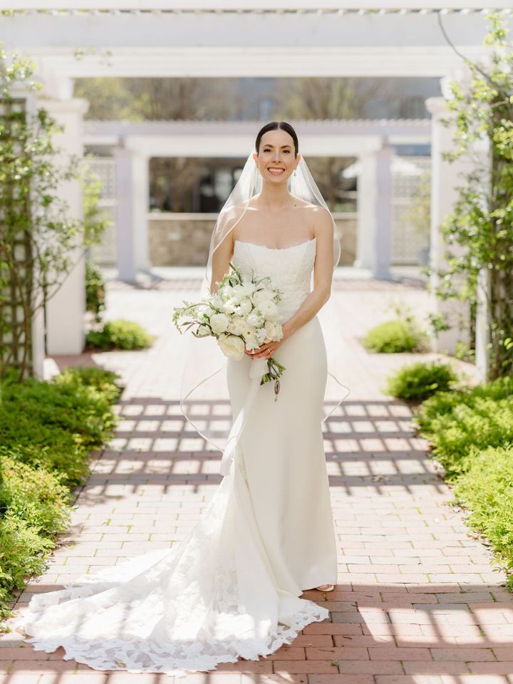Bride poses with white spring bouquet under white pergola.