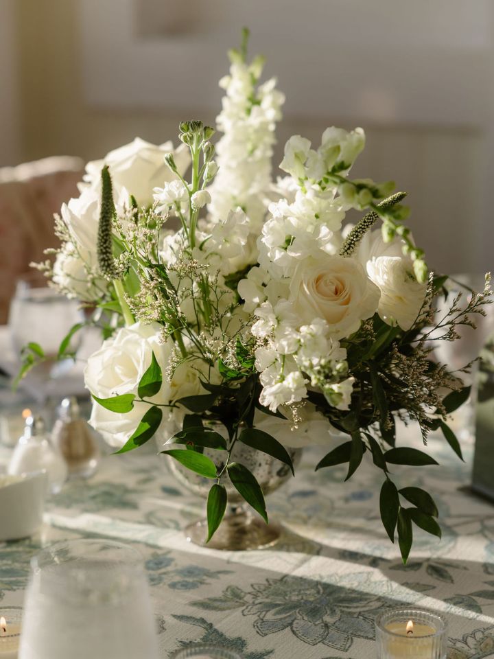 White flower arrangement in silver compote on patterned blue tablecloth.