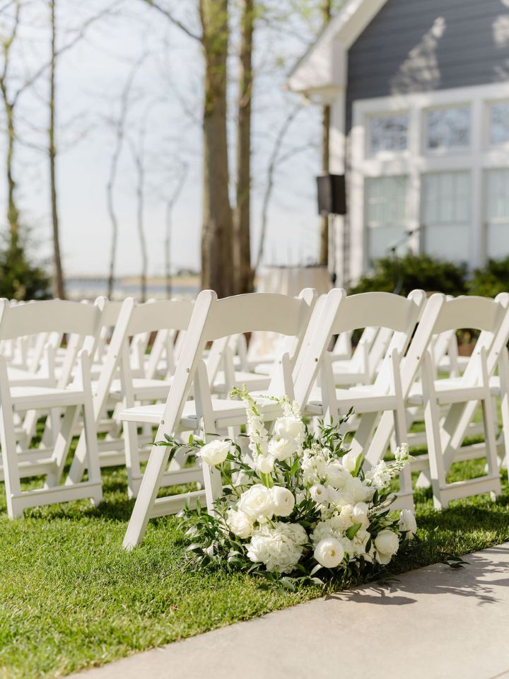 White folding chairs for an outdoor wedding ceremony with flower arrangement at base.