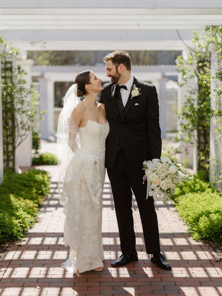 Bride and groom smile at each other under large white garden pergola.