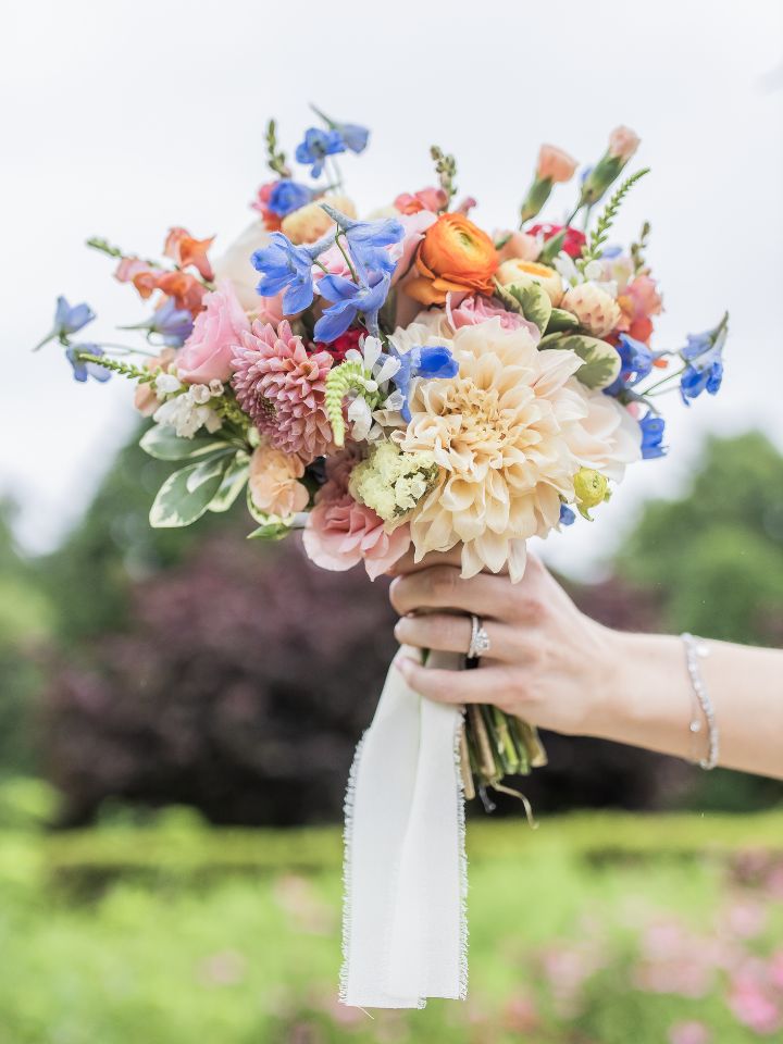 fairers bouquet Bride holds bouquet up to sky, full of light blue, peach, pale pink, and pops of orange flowers.