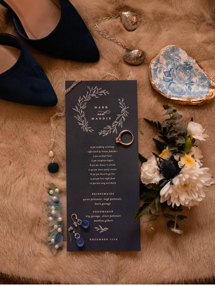 Flat lay of winter wedding invitation, oyster shell, jewelry, fur stole, and flower boutonniere.